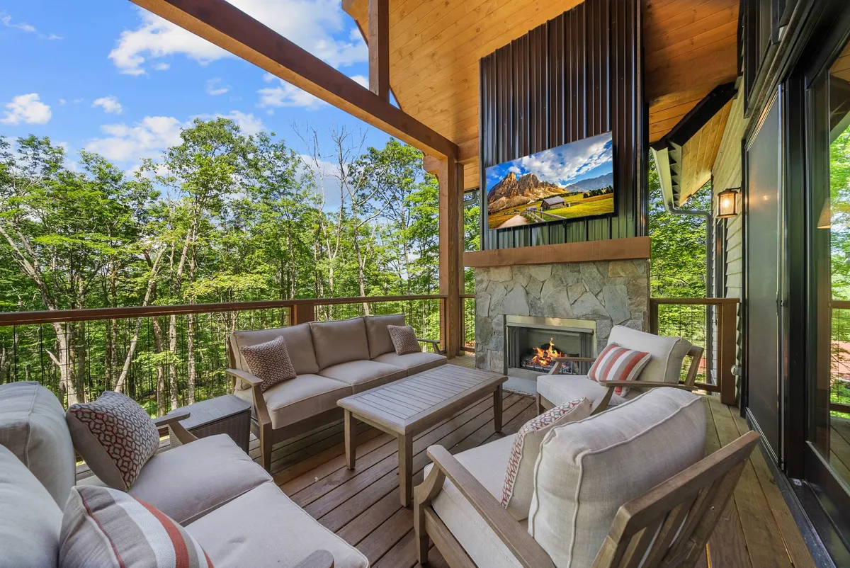 A spacious covered deck featuring a comfortable seating arrangement, a stone fireplace, a mounted TV, and surrounding wooden elements. Greenery and mountain views are visible in the background, blending indoor comfort with outdoor beauty.