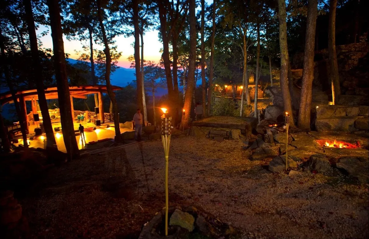 An inviting outdoor seating area with torch lights, a fire pit, Adirondack-style chairs, and panoramic mountain views in the distance. The glow of the fire and the warm lighting create a cozy, magical atmosphere for evening relaxation.