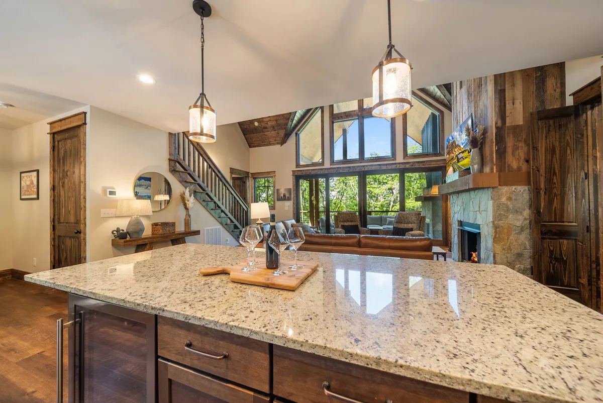 A luminous, welcoming interior featuring a granite kitchen island, large windows that frame scenic outdoor views, a dramatic stone fireplace, and a cozy leather sofa. The kitchen is integrated with the living space, encouraging natural flow and family interactions.