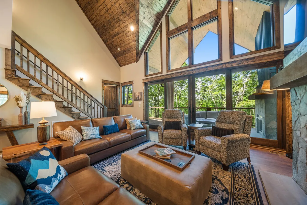 A bright, spacious interior with large windows flooding the room with natural light, cozy seating, a wooden staircase, and a fireplace. The views through the windows emphasize the ever-changing mountain landscape and seasonal transitions, tying the indoors to outdoor experiences.