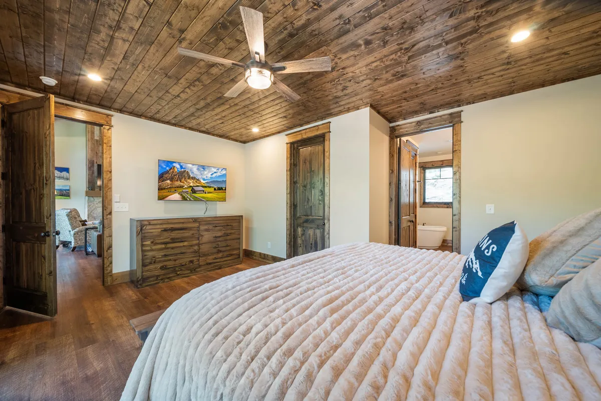 A comfortable bedroom with a bed dressed in a plush blanket, wooden furniture, a television, and a ceiling fan. The serene setting encourages relaxation, whether enjoying a movie or a quiet morning.