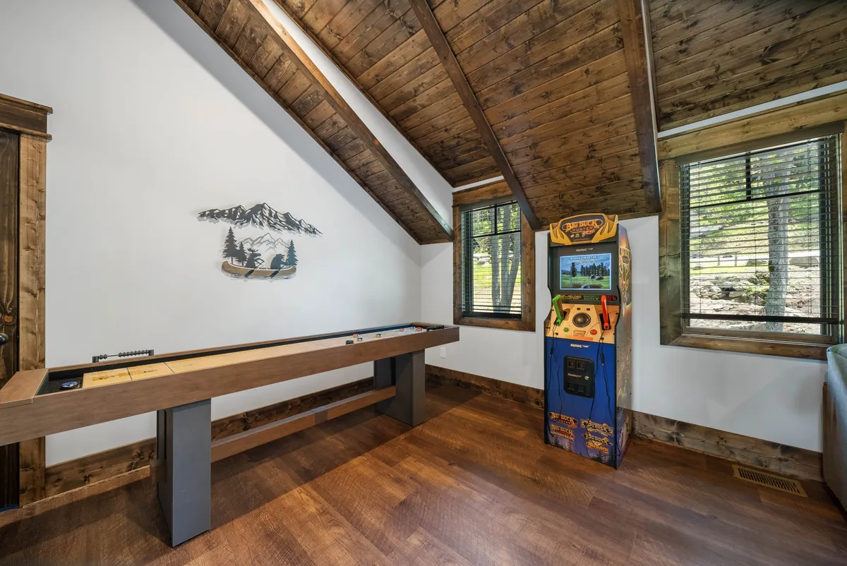 A lively loft space featuring a shuffleboard table and a classic arcade game machine, with mountain-themed wall art in the background. The area is open and well-lit, evoking a sense of playful energy and social interaction.