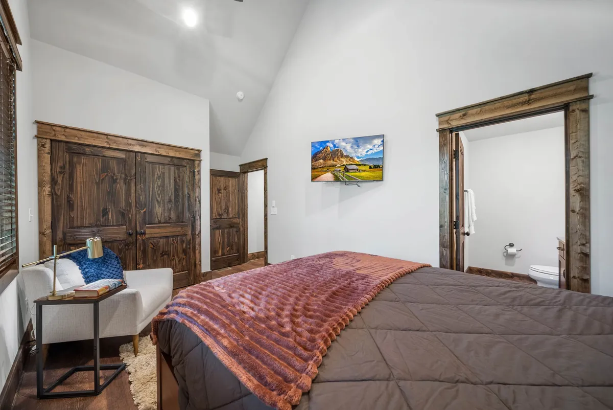 A spacious bedroom featuring a bed with a plush blanket, an armchair beside a side table, a TV mounted on the wall, and wooden closet doors. The room is filled with natural light and offers several cozy corners for relaxation.