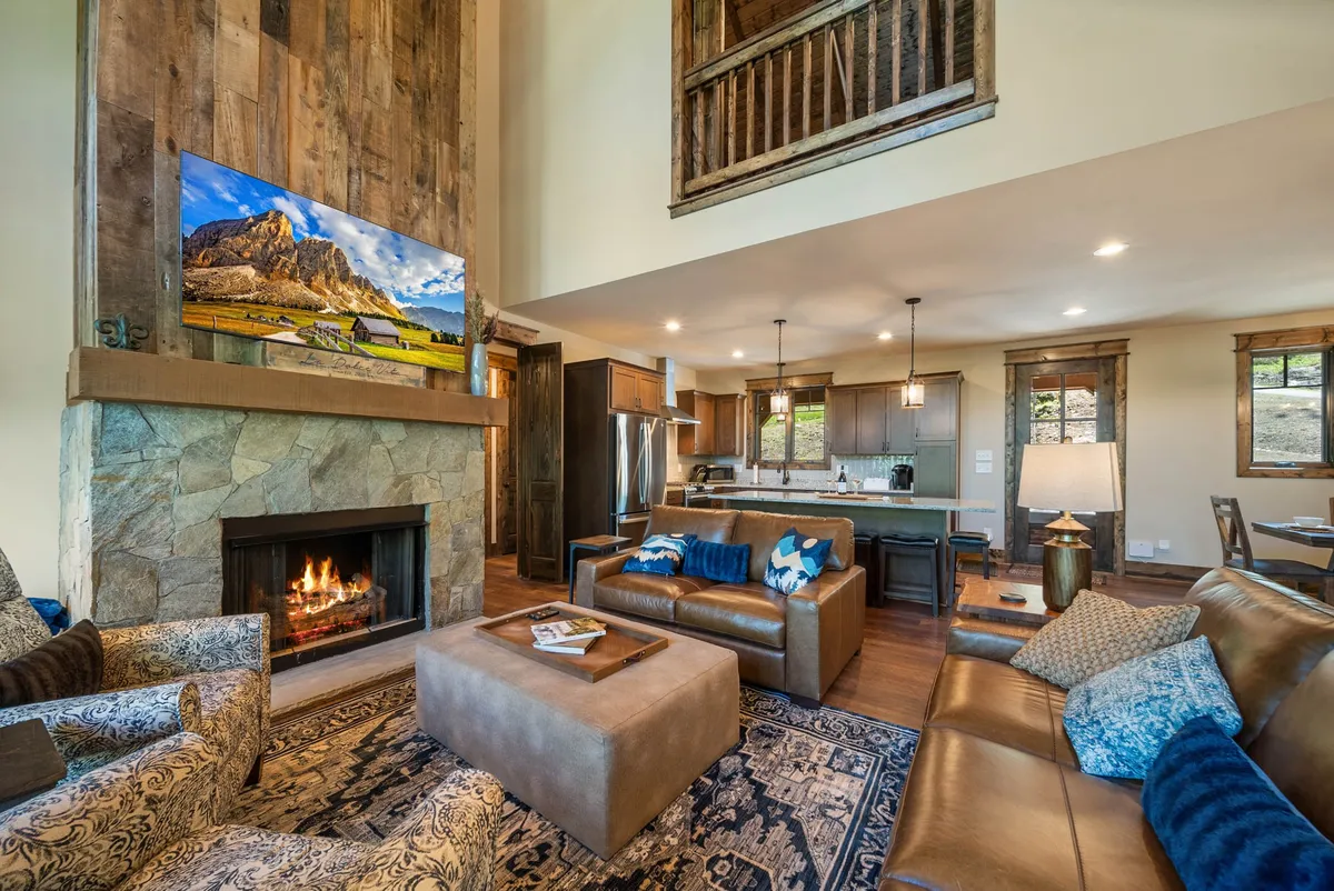 A warm, open-concept living area with a prominent stone fireplace, comfortable seating arranged for conversation, a television, and the kitchen area visible in the background. Large windows flood the space with natural light and offer glimpses of mountain views, while modern yet cozy decor ties the space together.