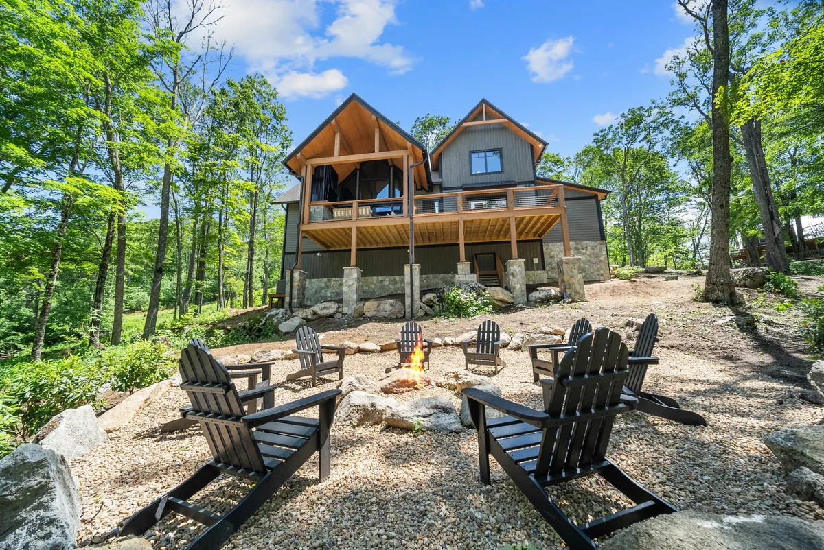 The image shows the warm exterior of a wooden mountain cabin nestled among tall trees, with a glowing fire pit encircled by Adirondack chairs in the foreground. The firelight creates a cozy, inviting atmosphere as dusk settles in, hinting at evenings spent sharing stories, roasting marshmallows, and enjoying the mountain air.