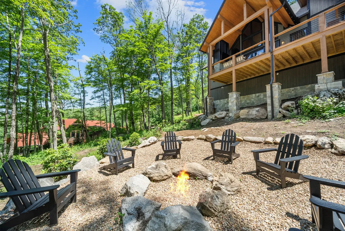 A classic firepit area with Adirondack chairs arranged in a circle, set against a backdrop of tall trees and the exterior of the house. The setting feels cozy and ideal for group gatherings or quiet reflection.